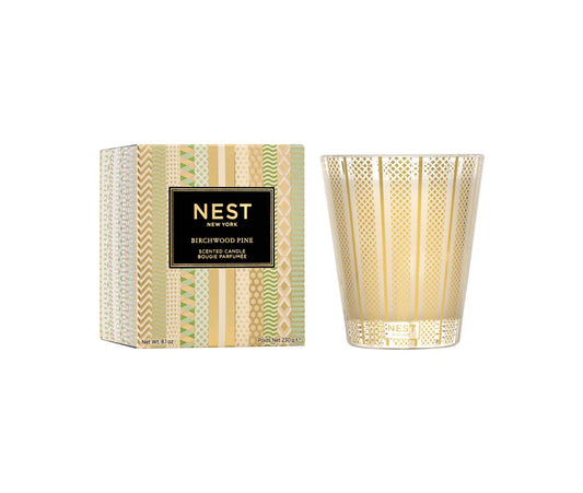 A clear glass candle with a geometric pattern and a gold label that reads 'NEST' and 'Birchwood Pine Classic Candle'.